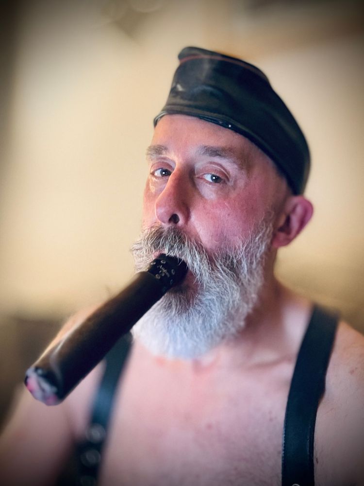 Nigel, white bearded, wearing leather suspenders and black leather forage cap  is bare chested, and smoking an asylum 8x80 cigar 