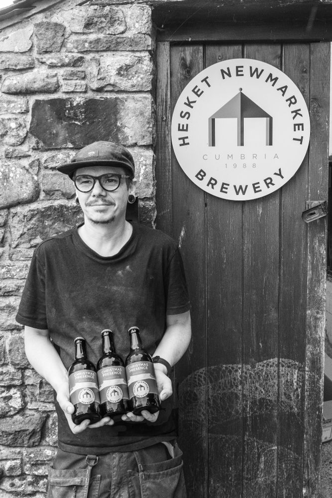A black and white photo of Shane, head brewer of Hesket Newmarket brewery, holding three bottles of a Christmas porter