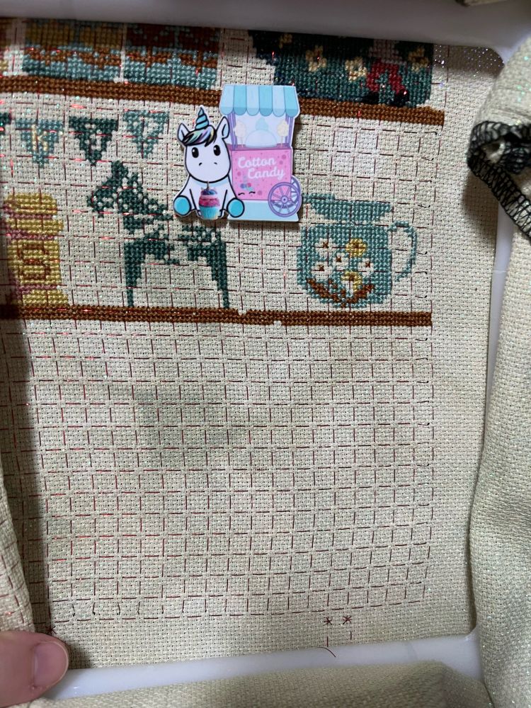 A section of cross stitch still in progress. 

The right side of the 2nd shelf crosses the rest of the pattern. There is a profile of a horse and there is a blue-green pitcher with partially colored in flowers. 

The needle minder is off a unicorn sitting in front of a cotton candy cart. 
