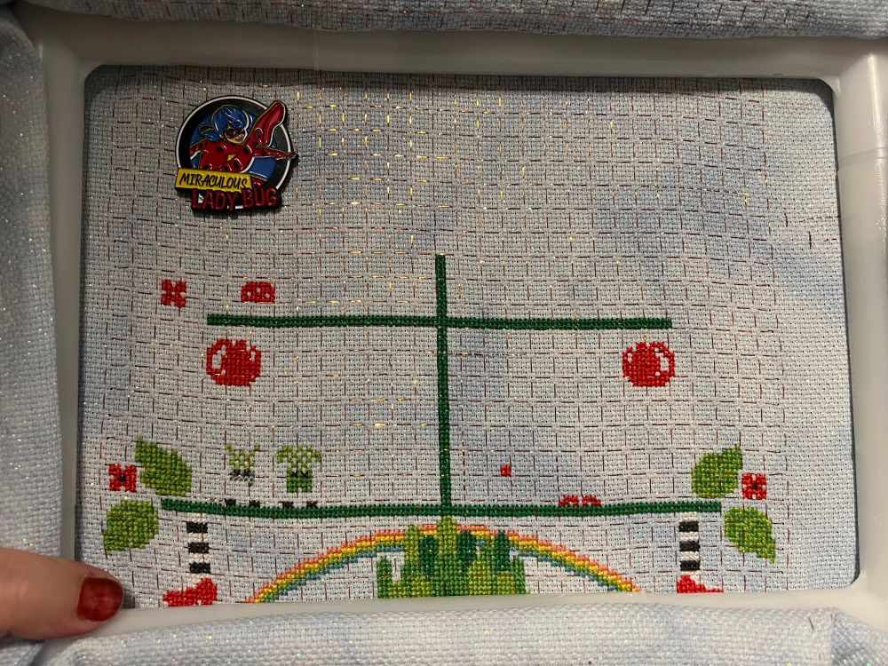 In-progress capture of The Wizard of Oz Tree by tiny modernist cross stitch. Tucked into the bottom of the Q-snap, you can see a rainbow over the towers of the Emerald City. On either side of the rainbow, striped stockings with red shoes hang down from a tree branch. Each side of the branch also sports some leaves and a red flower. Some red globes float near the top of the middle shelf. 

Needle minder is the superhero, Ladybug, doing a flying kick. The text reads Miraculous Ladybug. 