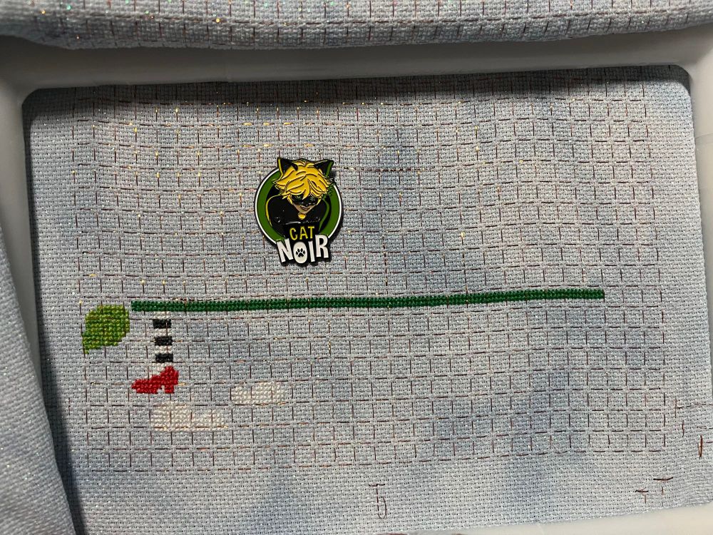 A leaf, a line across, and a stripy stocking with a ruby slipper. Two fluffy clouds hang underneath the shoe. 

Today’s needle minder is an image of Cat Noir’s head grinning. Under his face are the words Cat Noir dress 