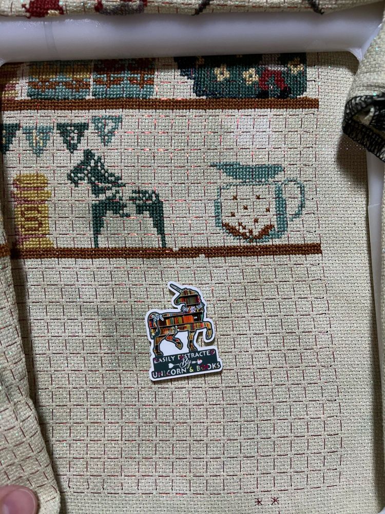 A section of cross stitch still in progress. 

The right side of the 2nd shelf finally crosses the rest of the pattern. There is a profile of a horse and an outline of a pitcher. 

The needle minder is an outline of a unicorn, around some bookshelves. The caption reads, “Easily distracted by unicorns and books”.