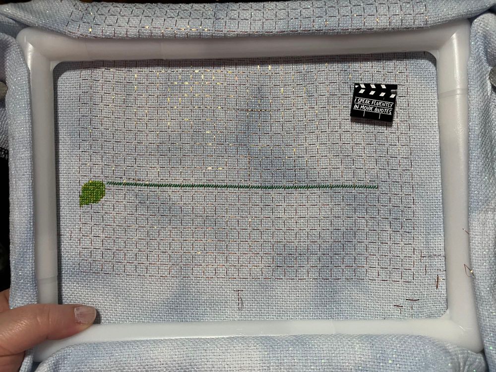 Gridded and mottled Aida fabric in a q-snap frame. There is a leaf on the left, and a line going across the middle of the fabric. 

The needle minder is a movie clapboard that says, “I speak fluently in movie quotes”.