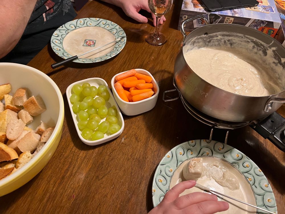 Cheeze fondue with carrots, grapes and a bowl of baguette cubes. There’s also a plate with Cheeze covered bread waiting to be eaten. 