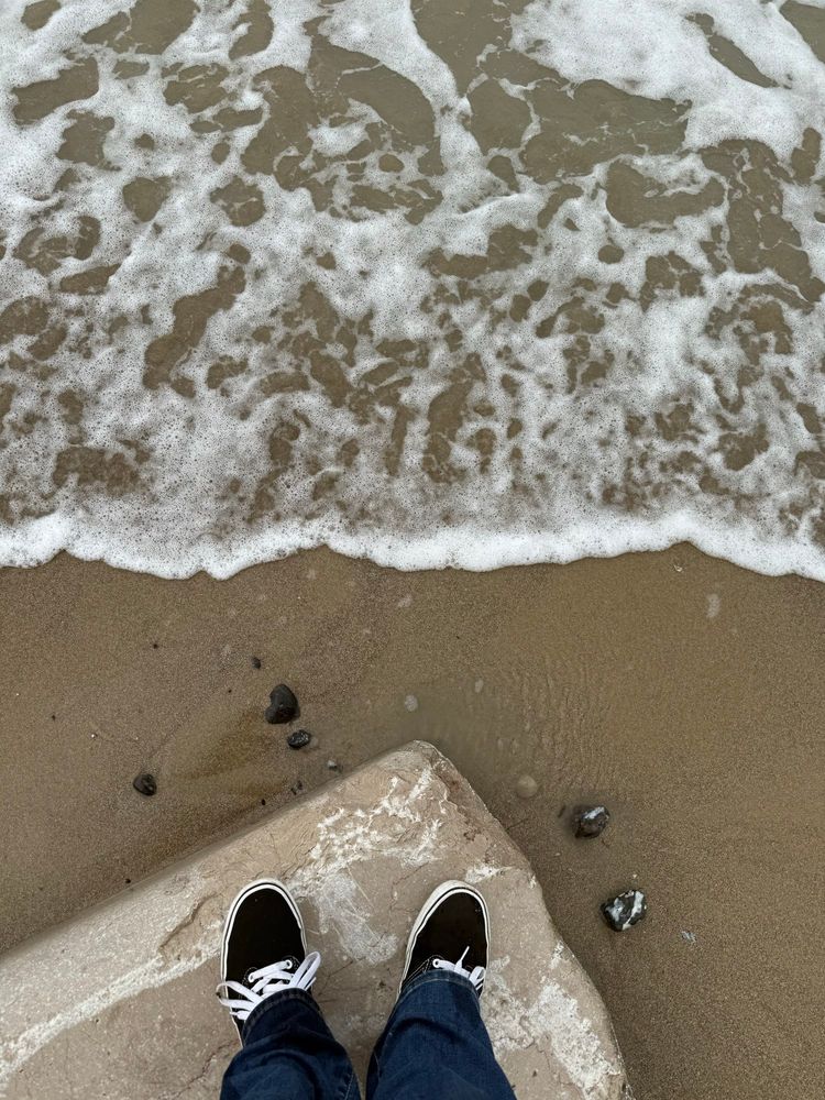 Füße in schwarz-weißen Vans auf einem hohen Stein, Sandstrand und eine kleine Welle ist zu sehen. 