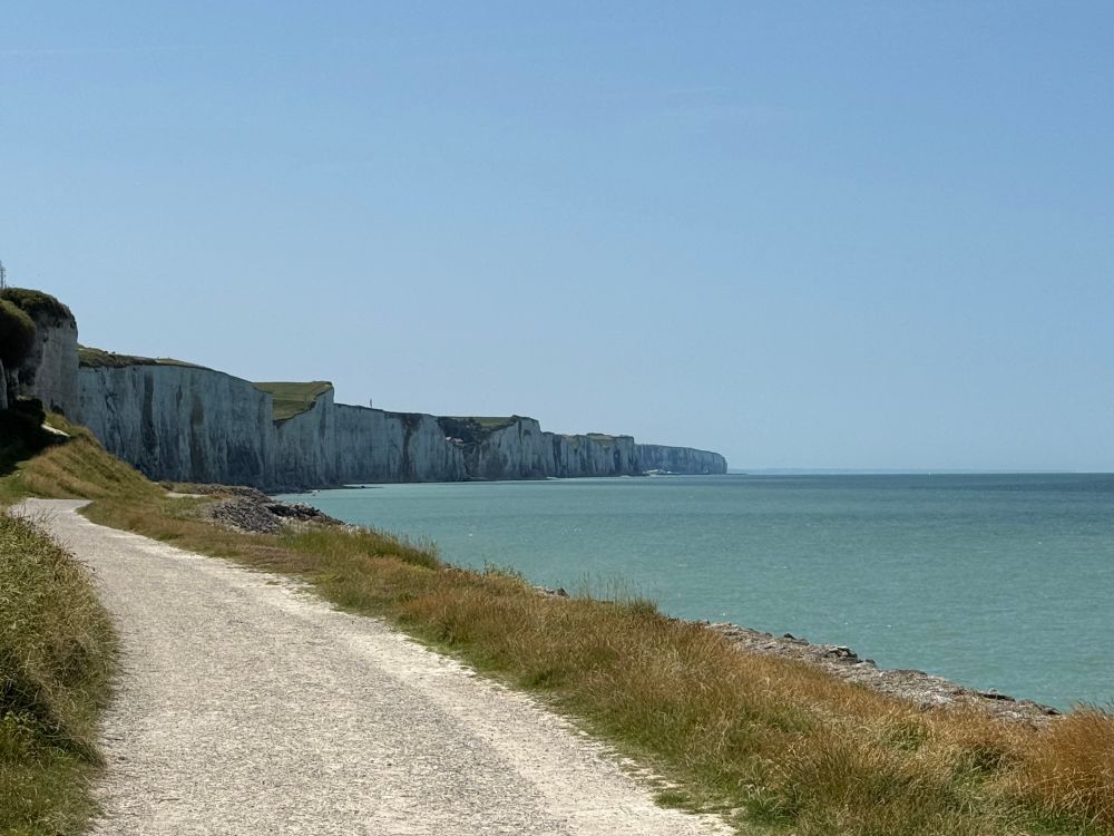 Zu sehen ist ein Weg am Meer. Strahlend blauer Himmel, ruhiges Meer und am linken Rand sind die Steilklippen zu sehen. 