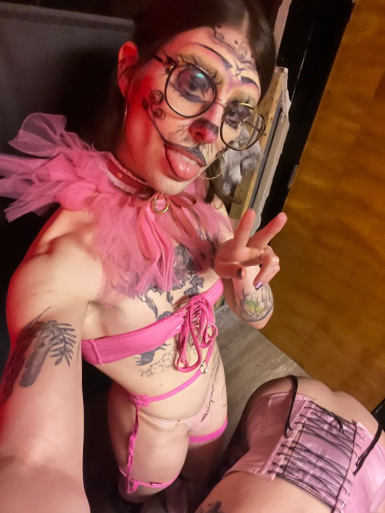bunny clown woobie in her pink get up,m from the first pic, but now she's standing, throwing a peace sign, tongue out, but now, she has a hot bimbo in a pink corset on her hands and knees. don't you wish you could be her 🫢