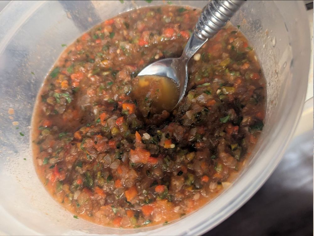 A plastic bowl full of salsa, with a spoon in it.  The salsa is quite chunky, and green, red, white, and shades in between are visible.