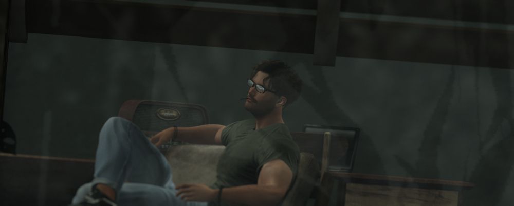 Digital representation of a man sitting in a chair. He is wearing a green t-shirt, faded denim jeans and Vans-style shoes. He has one leg up and is looking off into the distance a bit. We see the reflection of light in his glasses.  We are seeing him through a window; there is a hint of window frame and partially-drawn window blinds in the shot, as well as reflections/shadows in the hazy glass. 