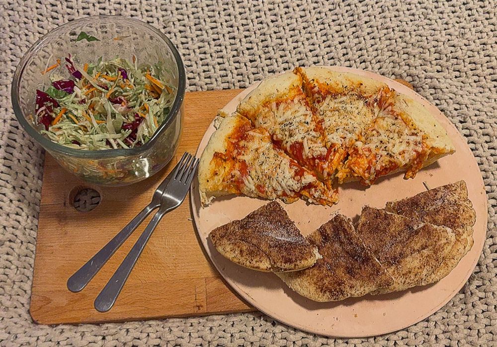 a beige blanket with a wooden cutting board. on top of the cutting board is a beige pizza stone (with four slices of pizza and four slices of cinnamon sticks), two silver forks, and a clear glass bowl filled with shredded salad. 