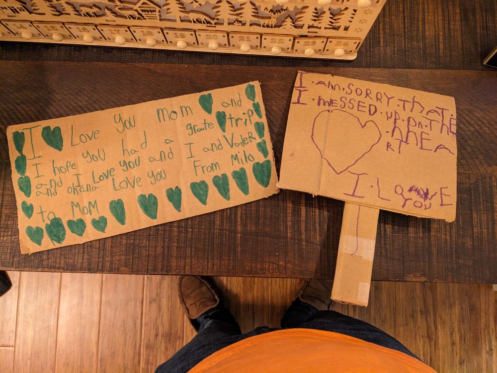 Two marker on cardboard signs. The one on the left reads "I love you Mom and I hope you had a grate trip and I love you and I and Vader and Ohana love you. From Milo, to Mom". The one on the right has a large, lopsided heart and reads "I am sorry that I messed up on the heart. I love you."