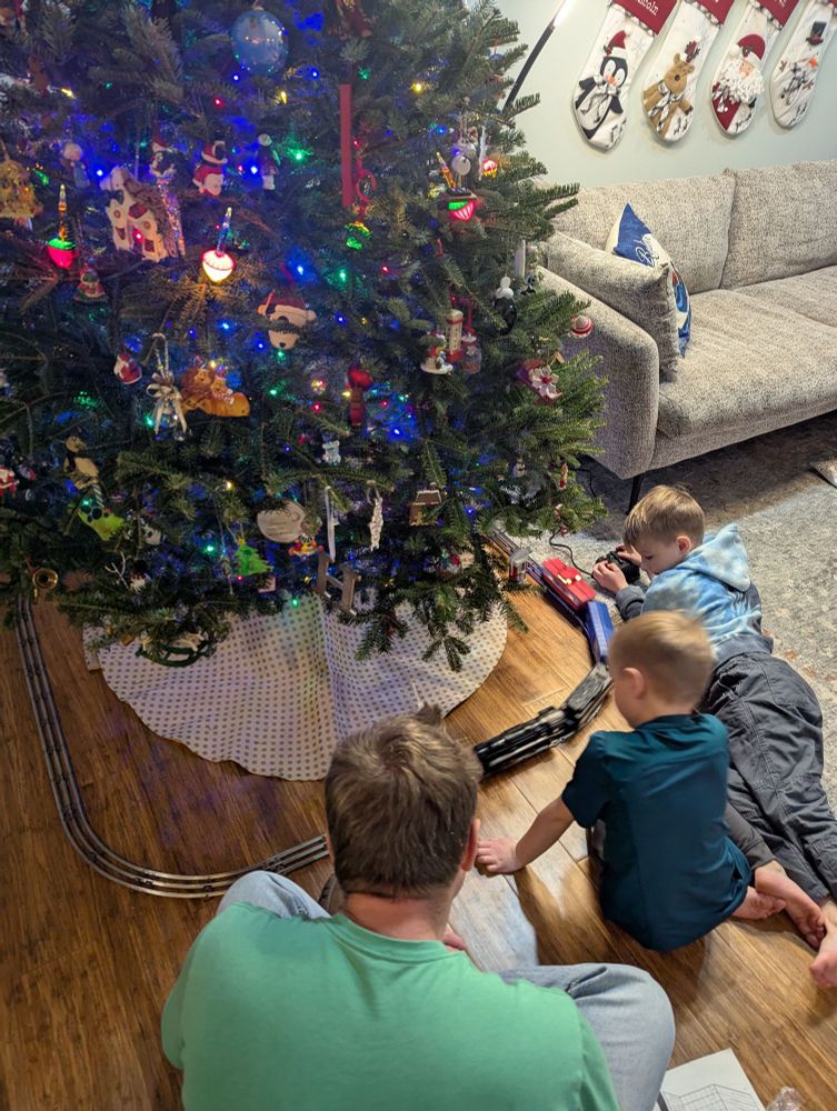 A Christmas tree with a Lionel train set around the base. Two boys and I sit on the floor playing with the train. 