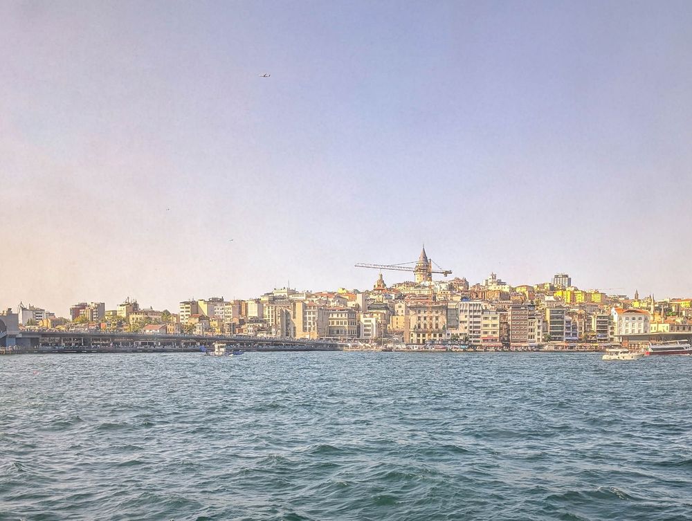 Galata bridge, tower, and seaside in the hazy light of mid afternoon sun, seen across the choppy water of the Bosphorus.