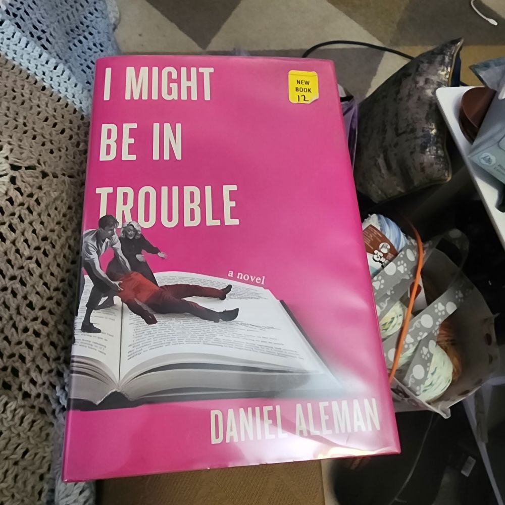 Cover image of I Might Be in Trouble by Daniel Aleman, a pink cover with a man and woman dragging a corpse across the pages of an open book 