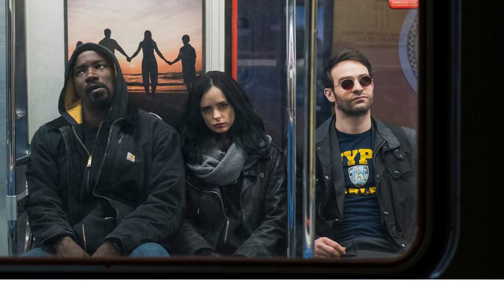 Imagem da série com o Luke Cage, Jessica Jones e Matt Murdock sentados no banco de um Metrô