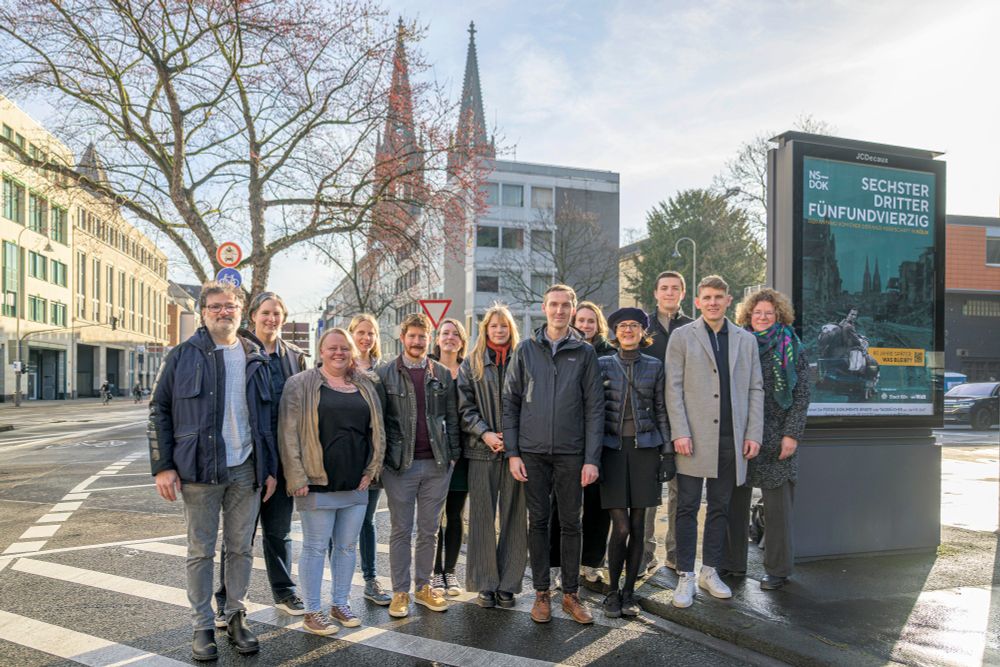 Gemeinsam posiert eine Gruppe Menschen neben einer digitalen Stadtinformationsanlage, auf der die Kampagne des NS-Dokumentationszentrums von Köln zu sehen ist. Im Hintergrund sind die zwei Türme des Kölner Doms zu sehen.