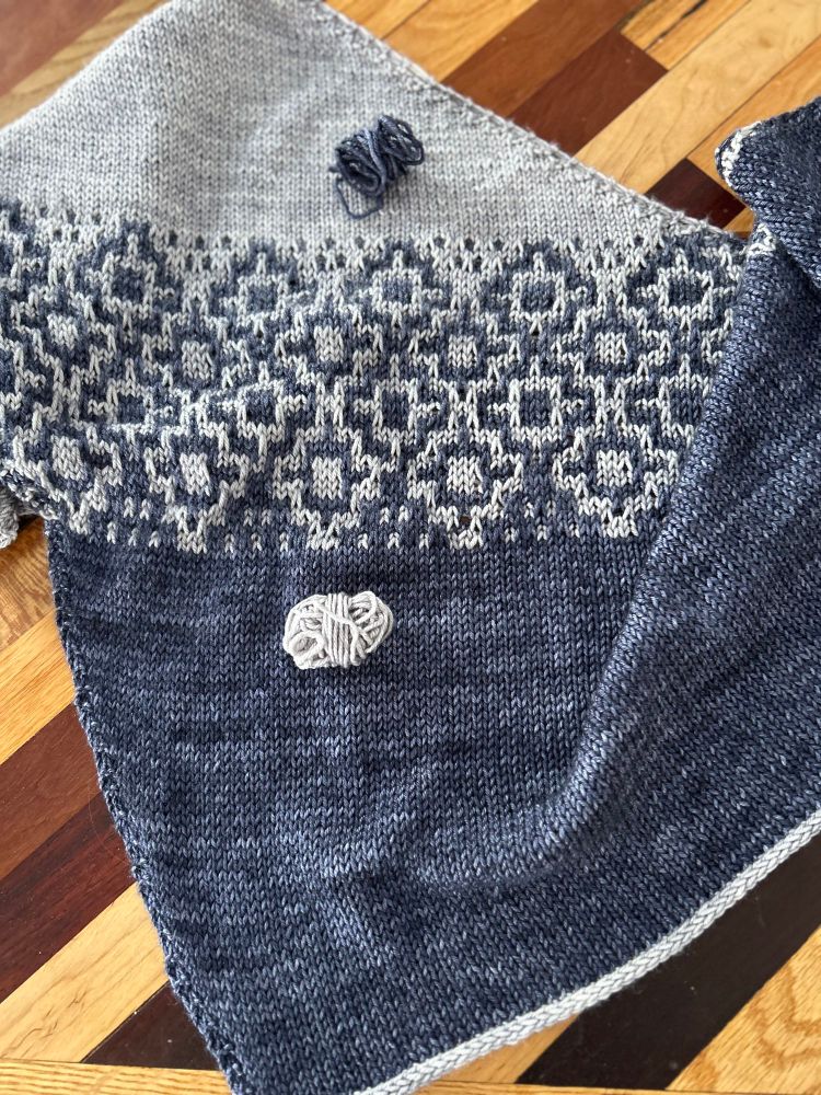 A close up of a shawl with mosaic color work in grey and blue. It also has the leftover balls of yarn 🧶 showing almost all was used. 