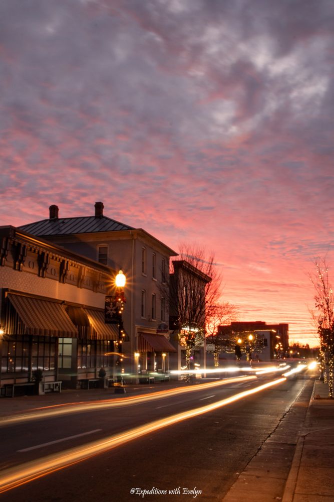 Clouds fill the sky and transition from purple to pink to peach to yellow during a winter sunset behind the buildings of a historic downtown area.  The street is decorated with holiday lights and in the center of the street, a long exposure effect has been used to create lines from passing cars.