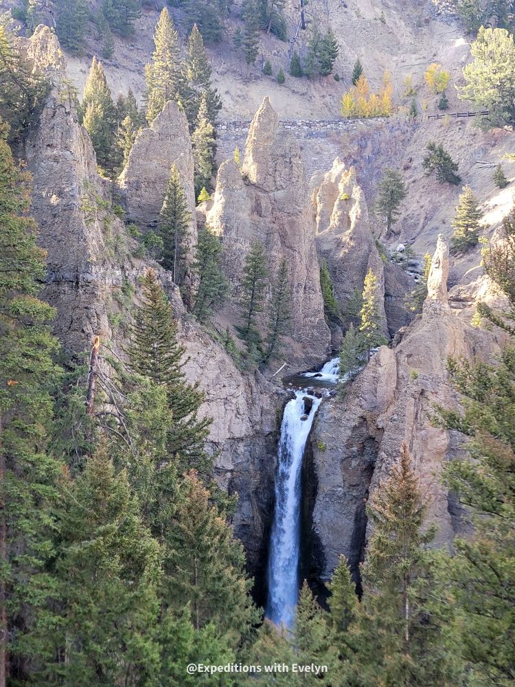 A streamlined waterfall crashes straight down from orange rocks to a pool behind green pine trees. The fading sun illuminates the tops of the rocks.