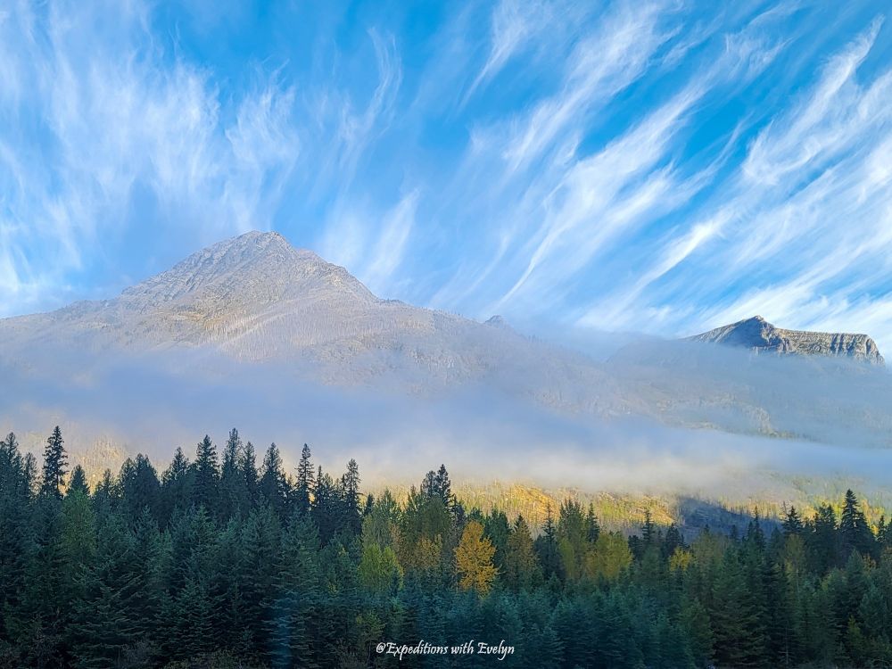 Wispy white clouds decorate the sky above mountains shrouded by thin fog.  Below the mist, golden trees amidst the greenery indicate the onset of fall.