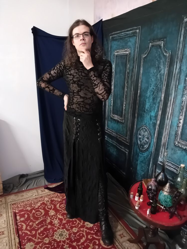 Goth man wearing a stringy black see-through top and black long skirt holding a hand to his face and pulling a pondering pose.