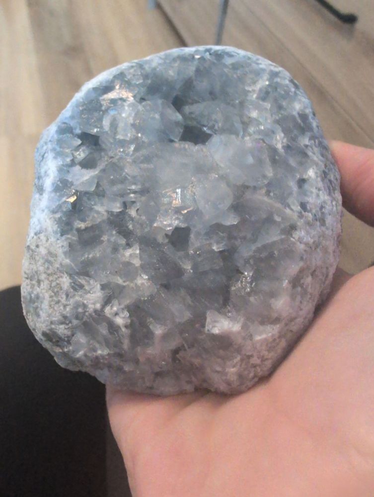 A hand holding a piece of celestite. It's a white geode on the outside, with the inner piece being full of pale blue crystals. 