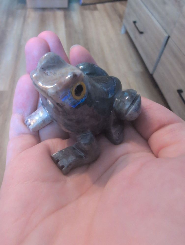 A hand holding a frog carved out of soapstone. He's mostly grey, with cream coloured streaks here and there. He has yellow eyes. 