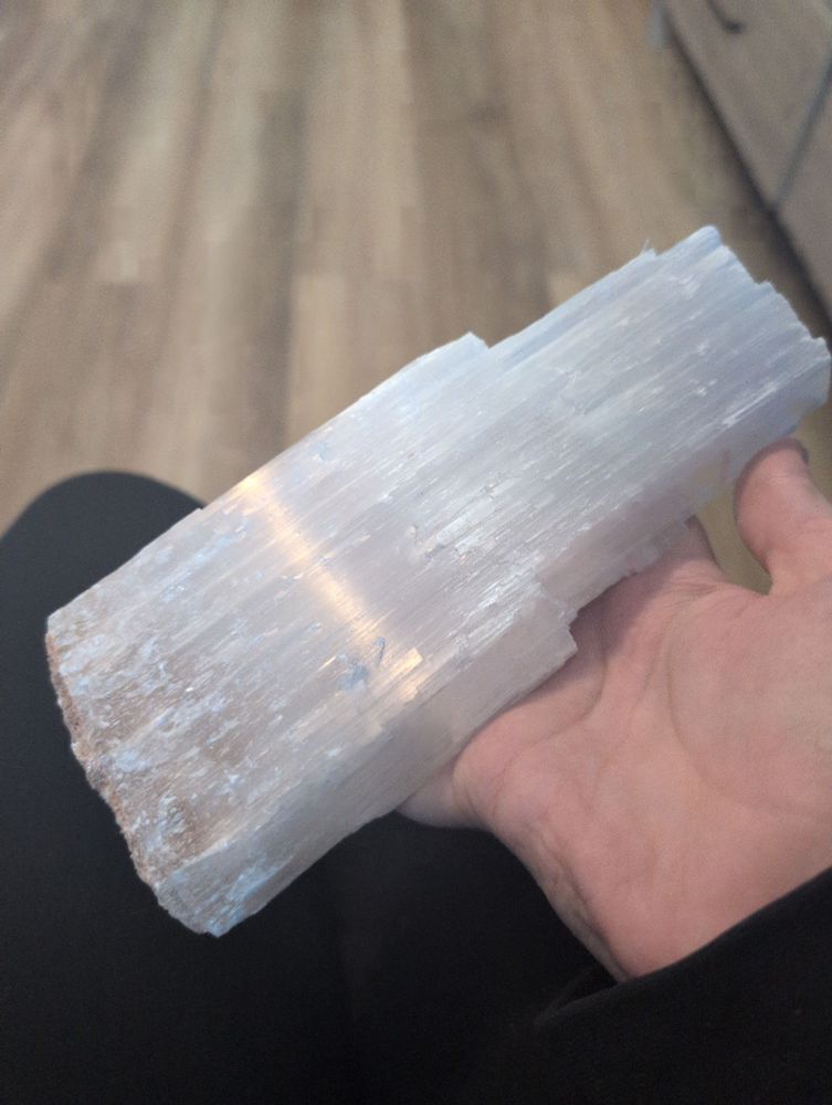 A hand holding a large piece of selenite. It's almost a pearl white color, with a texture reminiscent of a log of firewood. 