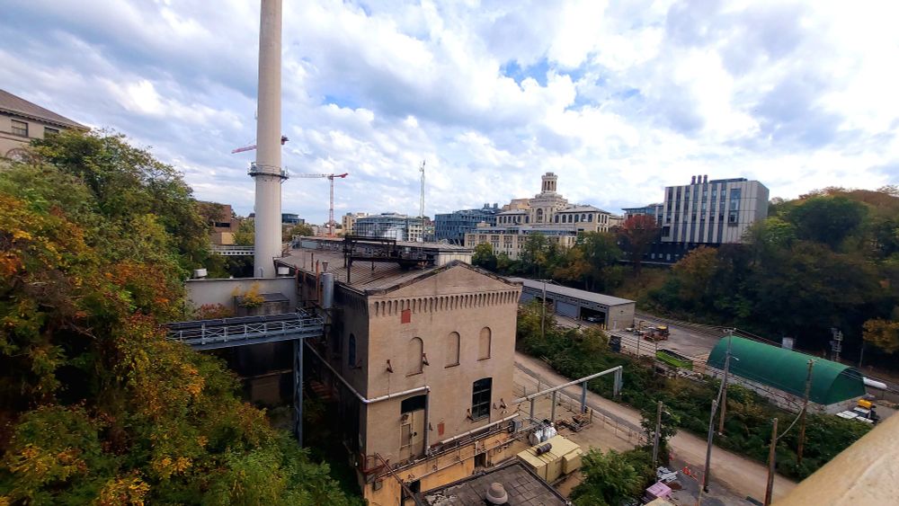 Concrete industrial building with a smokestack in a tree-lined valley with a few buildings on the far ridge and blue skies with white clouds in the background