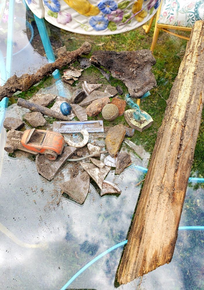 A pile of dirty odds and ends on a glass tabletop including a wooden stake, metal spike, red toy car, blue marble, and several random pieces of glass, plastic, and porcelain.