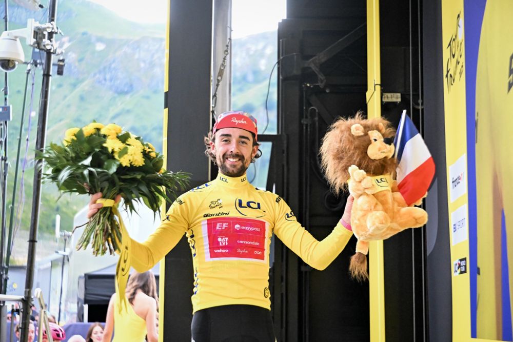 photo of a white man (ben healy) in pro cyclist attire. he is wearing a yellow jersey and holding a bouquet in one hand and a stuffed lion in the other. he is smiling 