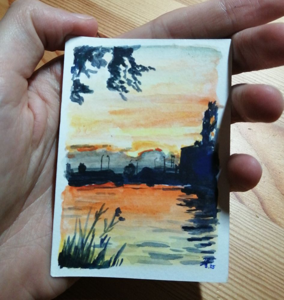 Kleine Aquarellskizze, die in der Hand gehalten wird. Darauf zu sehen in Orange und dunklen Blautönen ein Fluss bei Sonnenuntergang mit Gebäude- und Vegetationssilhouetten.