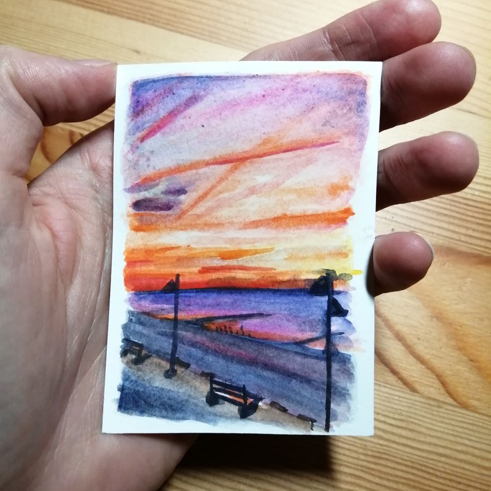 In der Hand gehaltenen kleine Aquarellskizze. In Orange, Violett und Blaugrau gehaltene Abendstimmung am Meer, im Vordergrund eine Bank und Laternen