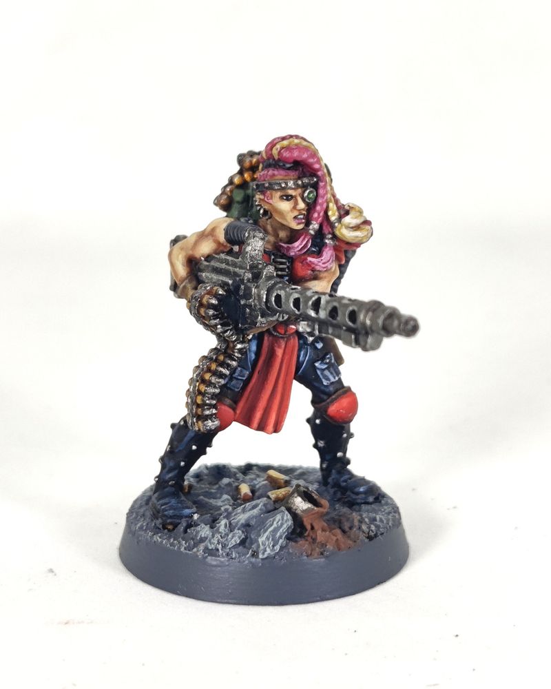 Photograph of a 1995-era Necromunda Escher Heavy miniature on a white background. The model is a muscular amazon-like woman holding a large, WW2-esque belt-fed machine gun, specifically a heavy stubber. The woman has a multitude of pink and blonde braids, a bionic targeter eye and red armour with studded black leather chaps over faded blue jeans. She is standing on a grey, rocky base with broken pipes and fallen shell casings.