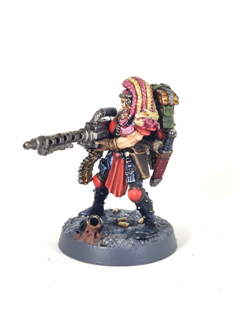 Photograph of a 1995-era Necromunda Escher Heavy miniature on a white background. The model is a muscular amazon-like woman holding a large, WW2-esque belt-fed machine gun, specifically a heavy stubber. The woman has a multitude of pink and blonde braids, a bionic targeter eye and red armour with studded black leather chaps over faded blue jeans. She is standing on a grey, rocky base with broken pipes and fallen shell casings.