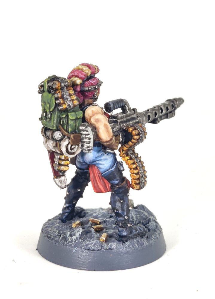 Photograph of a 1995-era Necromunda Escher Heavy miniature on a white background. The model is a muscular amazon-like woman holding a large, WW2-esque belt-fed machine gun, specifically a heavy stubber. The woman has a multitude of pink and blonde braids, a bionic targeter eye and red armour with studded black leather chaps over faded blue jeans. She is standing on a grey, rocky base with broken pipes and fallen shell casings.
