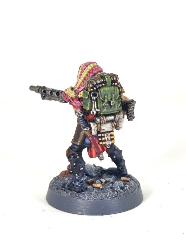 Photograph of a 1995-era Necromunda Escher Heavy miniature on a white background. The model is a muscular amazon-like woman holding a large, WW2-esque belt-fed machine gun, specifically a heavy stubber. The woman has a multitude of pink and blonde braids, a bionic targeter eye and red armour with studded black leather chaps over faded blue jeans. She is standing on a grey, rocky base with broken pipes and fallen shell casings.