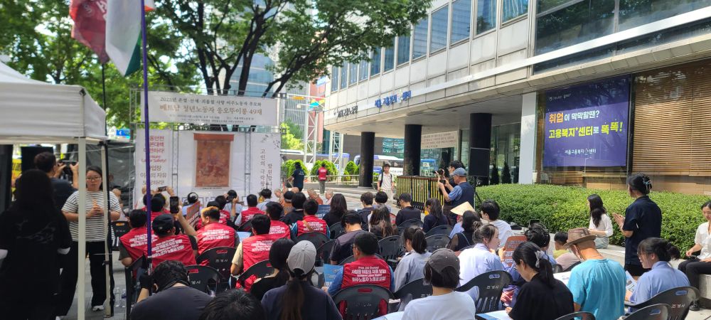 서울지방고용노동청 앞에 이천이십오년 온열·산재·괴롭힘 사망 이주 노동자 추모 및 베트남 청년 노동자 응오주이롱 사십구재를 위한 단이 마련되어 있으며 그 앞으로 사람들이 의자에 앉아 있다.