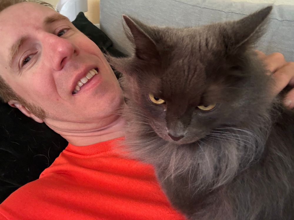 Bacho laying on couch wearing a bright orange shirt with a stupid from. His beautiful multicolored grey floofy cat, with an outrageous bib, is sitting on his upper chest looking nonchalant.