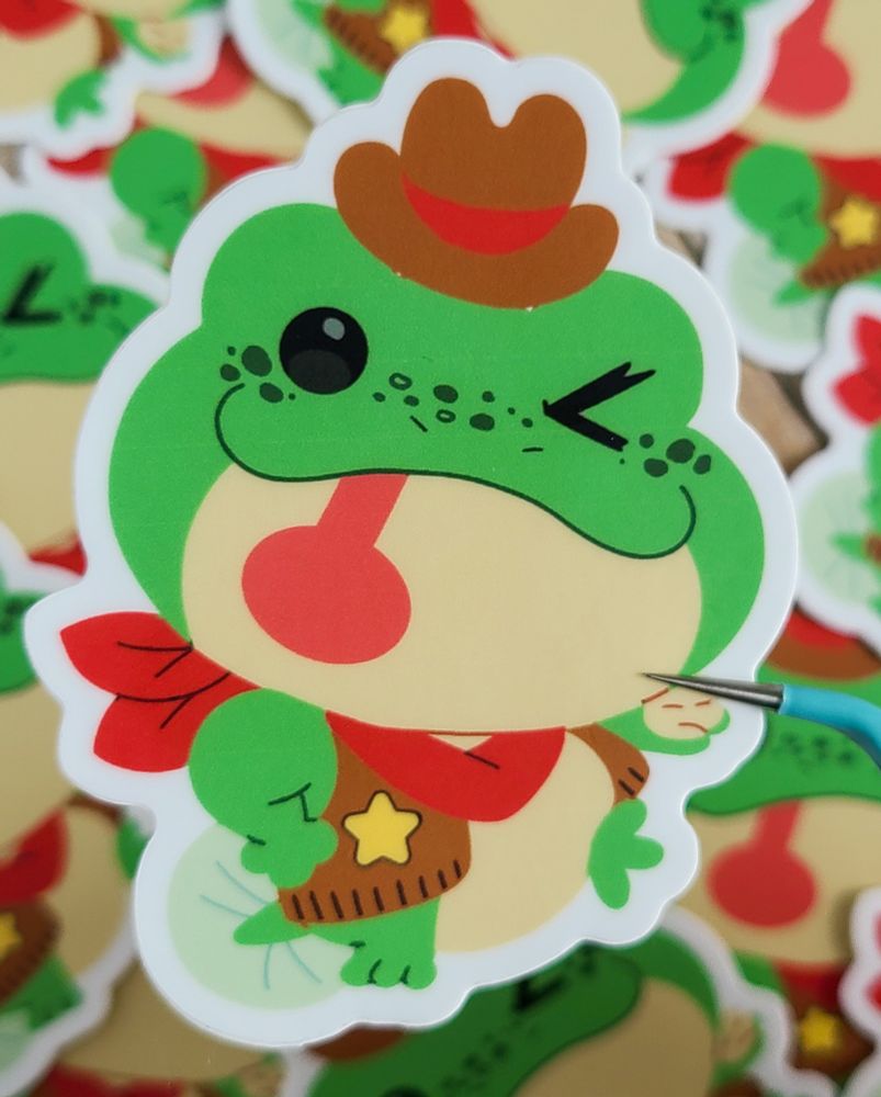 A sticker of a green frog dressed in a brown cowboy outfit with a red bandana.It is sticking out it's red tongue. 