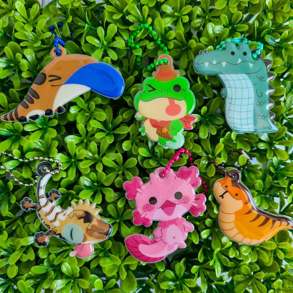 6 shiny puffy charms of various animals lie on a green bushy background. From top left to bottom right: A blue tongued skink, a frog, a crocodile, a crested gecko, an axolotl creature, and a short snake, the tsuchinoko. 