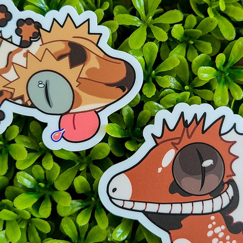 A close up of two stickers. Two brown, white, and black crested geckos lying on a green, leafy background. 