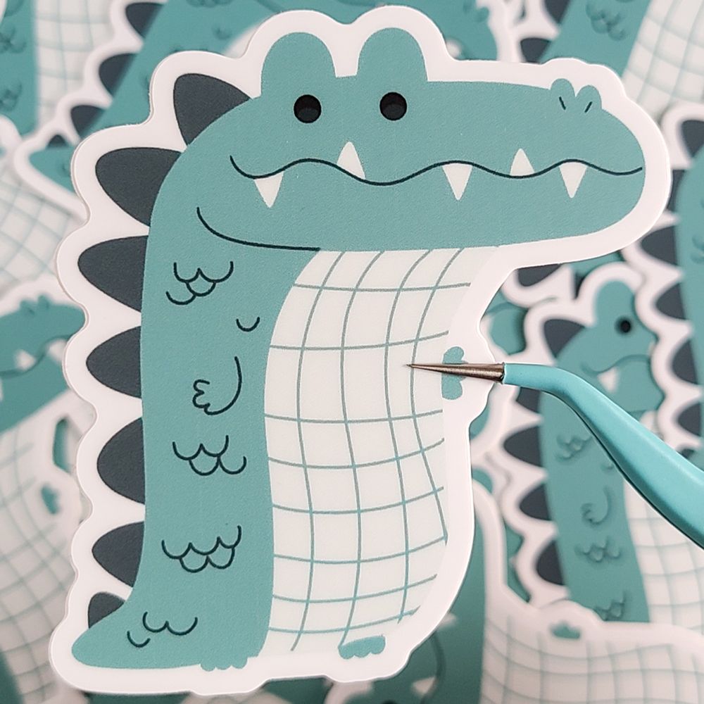A sticker of a long, green and white alligator / crocodile. It has a wiggly smile with teeth poking out. 