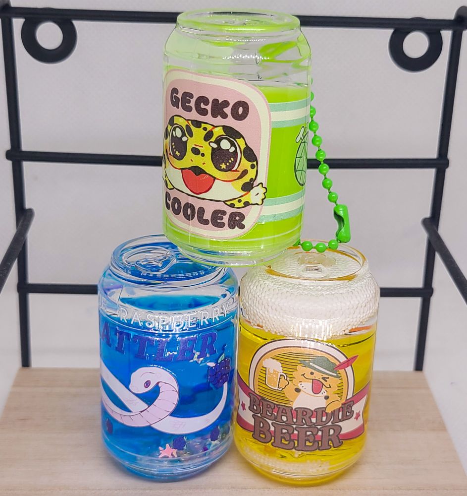 Three colorful soda can charms are stacked on top of each other. Green, blue and yellow cans with different reptiles on them. 