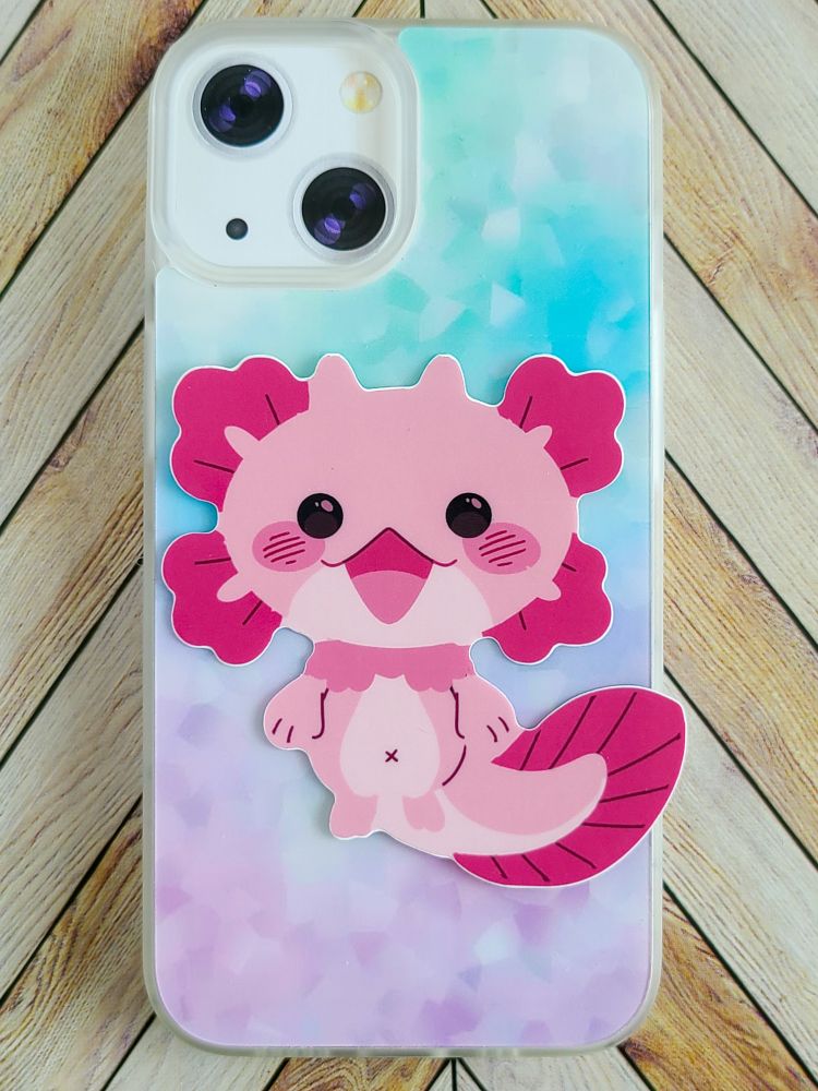 A sticker of a pink axolotl type creature sits on a Samsung phone case. It has beady black eyes and a big smile on its face. 