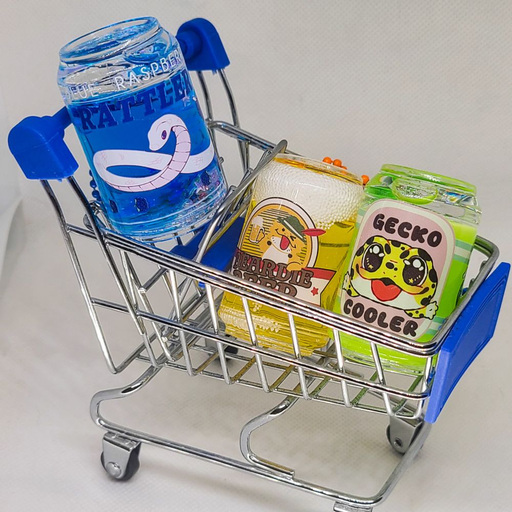 A toy shopping cart filled with three colorful soda charms. They are blue, yellow, and green. Each soda has a different reptile and design on them. The first is a white snake, the second is a bearded dragon with the phrase "beardie beer", and the last is a leopard gecko with the phrase "gecko cooler". 
