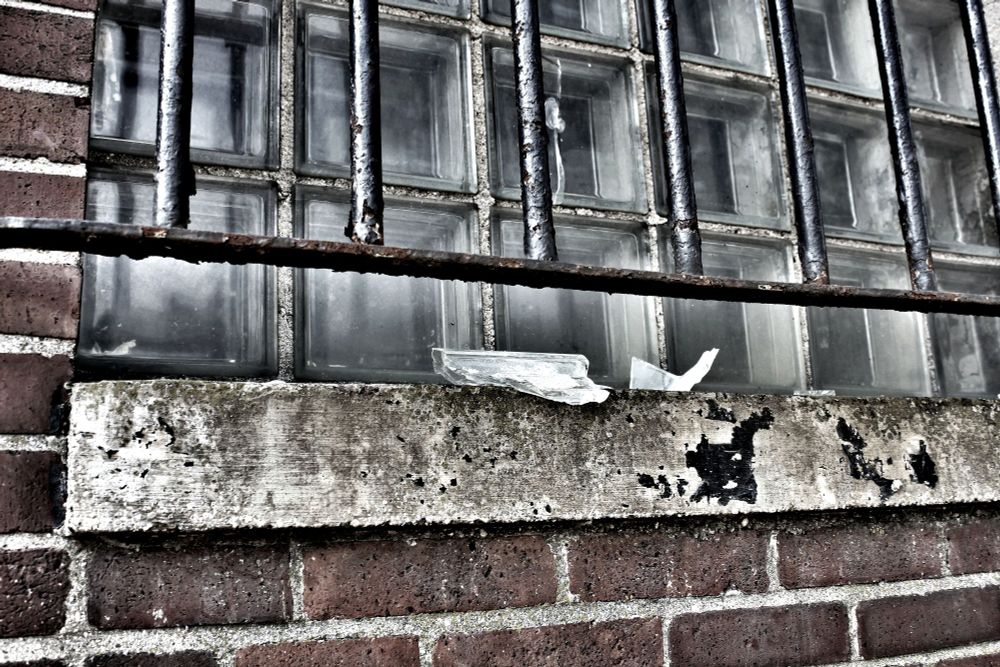 Slightly desaturated image of a block window, barred, in a red brick wall. Broken block glass sits on the sill, while rust eats away at the black paint on the bars. Old concrete and mortar is stained with age and probably mold, maybe some paint. Probably taken at an outbuilding at an active prison in Illinois or somewhere like that. 