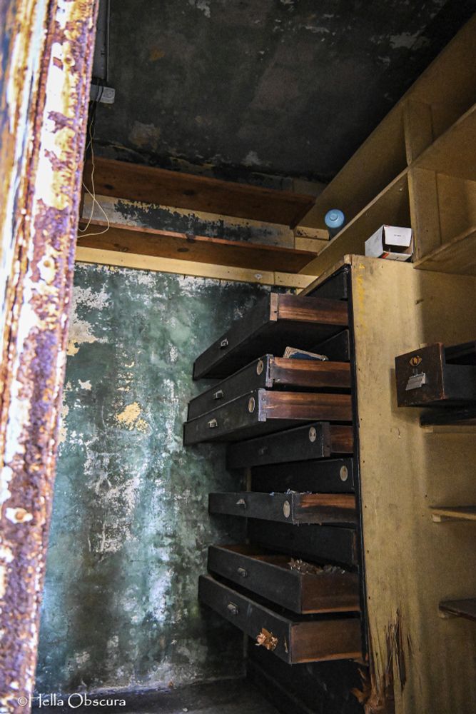 A photo of a chest of drawers, each drawer is open to varying degrees. It's in an old closet with built-in shelves, peeling green paint, and clearly abandoned. The drawers are wood, stained dark, but the sides of the dresser have been worn to their natural lighter color. A single metal drawer sits on a shelf next to the dresser, open about halfway, rusting and probably full of tetanus. 