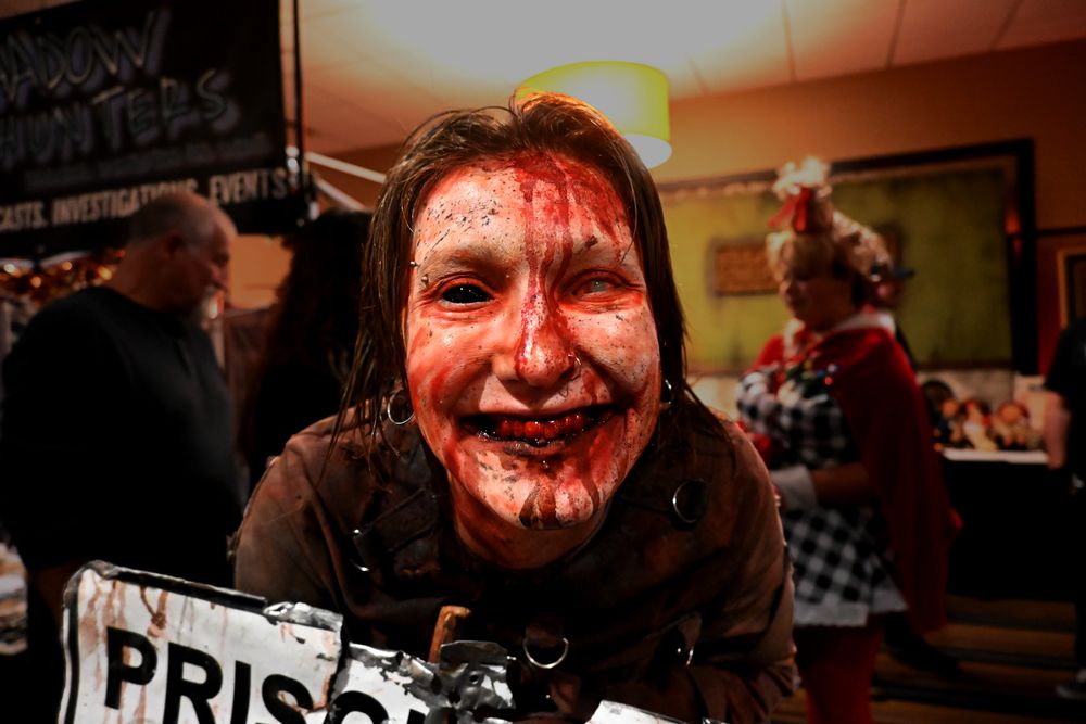 The haunt actor String Cheese poses at Days of the Dead in Chicago, 2024. She's in focus while the background is dimmer and unfocused. She has one black eye, and one white marble eye with blood streaking down her face. She holds a signpost, the sign reading Prison Area Do Not Pick Up Hitchhikers in reference to the derelict Illinois State Prison - Joliet. 