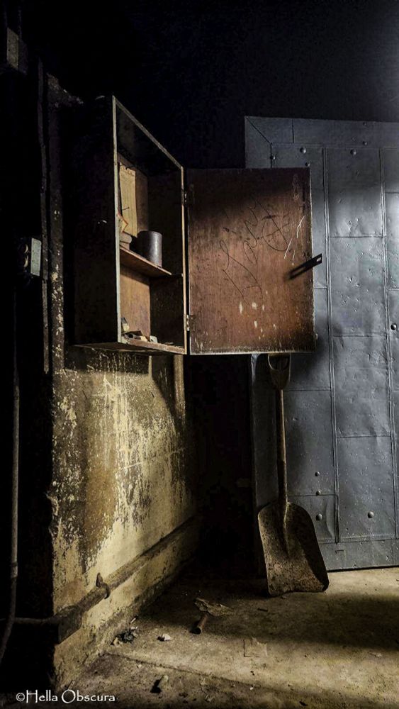 Photo in a dingy basement. We are inside a room, a large metal door to the right is open inward. We cannot see out of the room. A rusted coal shovel rests upright against the gray steel firedoor. A rusted metal cabinet hangs on the wall to the left, the cabinet door is open exposing a shallow shelf with various unidentified objects resting on it. Lighting is from the right, outside of this room, providing deep shadows